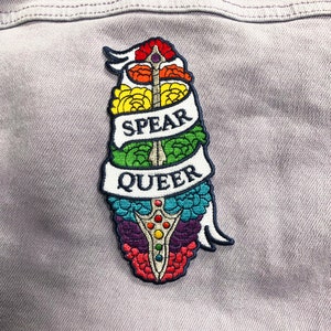 May include: An embroidered patch featuring a rainbow-coloured spear with the words "Spear Queer" on a white banner.