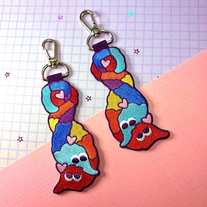 May include: Two colorful embroidered keychains with a whimsical design. Each keychain features a patchwork pattern in shades of red, blue, orange, purple, and yellow, with heart accents and a silver clasp. The keychains are attached to a purple strap.