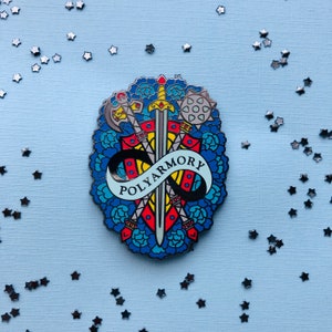 May include: A colourful enamel pin featuring a sword and other weapons crossed over each other, surrounded by flowers and the text "Polyarmory".