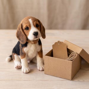 Puede incluir: Una figura de cachorro beagle de lana fieltrada con pelaje marrón, negro y blanco, que lleva un pequeño collar trenzado. Junto al cachorro hay una caja de regalo cuadrada de cartón marrón claro con una etiqueta y cordel.