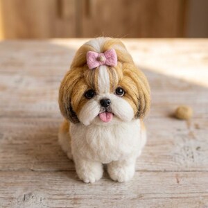 May include: A needle-felted Shih Tzu dog figurine with long, textured tan and white fur. The dog features a small pink bow on top of its head, dark round eyes, a black nose, and a small pink tongue visible between its white muzzle fur.