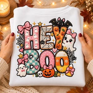 Puede incluir: Una sudadera blanca con un gráfico colorido que incluye el texto HEY BOO. El diseño presenta letras estampadas en rosa, turquesa y naranja decoradas con fantasmas, calabazas, arañas, telarañas, lazos y estrellas.