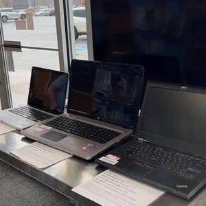 May include: Three laptops with black screens are arranged on a metal table. The laptops vary in size and color, including silver and black models. Below the table, a Guitar Hero II video game box and a game case are visible on a lower shelf.