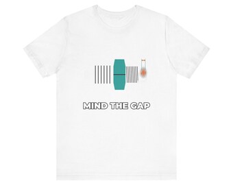 Könnte beinhalten: Ein weißes Kurzarm-T-Shirt mit einer Grafik aus einer türkisfarbenen geometrischen Form, flankiert von vertikalen schwarzen Linien und einem kleinen orangefarbenen Kreis. Darunter steht der Text MIND THE GAP in schwarzen Buchstaben.