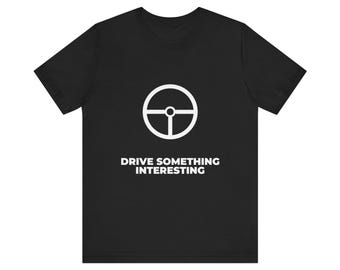 Könnte beinhalten: Ein schwarzes Kurzarm-T-Shirt mit einer weißen Grafik eines Lenkrads, das mittig auf der Brust platziert ist. Unter dem Lenkrad ist der Text DRIVE SOMETHING INTERESTING in einer fetten, weißen, serifenlosen Großbuchstabenschrift gedruckt.
