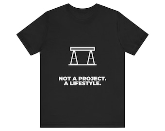 Könnte beinhalten: Ein schwarzes Kurzarm-T-Shirt mit einer weißen Strichzeichnung eines Sägebocks mittig auf der Brust. Unter der Grafik steht in weißen Großbuchstaben der Text: NOT A PROJECT. A LIFESTYLE.