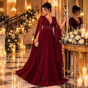 Può includere: Un abito lungo color bordeaux con un profondo scollo a V, maniche lunghe e fluide a mantella e una gonna plissettata. Il corpetto è decorato con piccole perline bianche lungo la vita e lo scollo, e l'abito è tenuto con una piccola pochette.