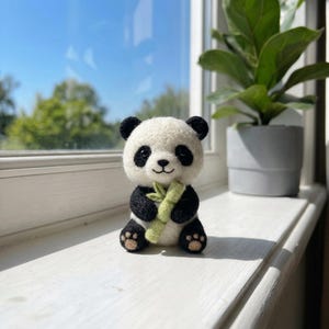 May include: A small, needle-felted panda figurine with a white and black body, black ears, and black eye patches. The panda has a smiling expression and is holding a light green felted bamboo stalk in its front paws, with small tan paw pads.