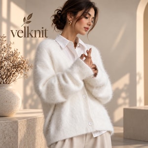May include: A white fuzzy V-neck cardigan with long sleeves and front buttons, worn over a white collared shirt. The text velknit is visible in the upper left corner, accompanied by a small leaf icon above the brand name.