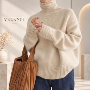 May include: A cream-colored, long-sleeved turtleneck sweater with a ribbed collar, cuffs, and hem. The text VELKNIT is visible in the upper left corner. The sweater is paired with dark blue denim jeans and a pleated brown leather handbag.