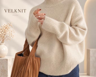 May include: A cream-colored, long-sleeved turtleneck sweater with a ribbed collar, cuffs, and hem. The text VELKNIT is visible in the upper left corner. The sweater is paired with dark blue denim jeans and a pleated brown leather handbag.