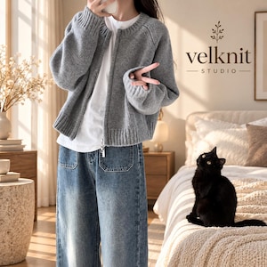May include: A grey knit zip-up cardigan with a round neckline and ribbed cuffs, worn over a white t-shirt and blue denim jeans with front pockets. The text 'velknit STUDIO' is visible in the upper right corner of the image.