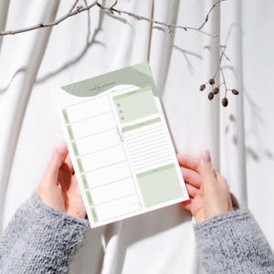 May include: A weekly planner sheet featuring a sage green and white color scheme with abstract shapes at the top. The layout includes sections for daily scheduling, a to-do list with checkboxes, a notes area, and a goals section with lines.