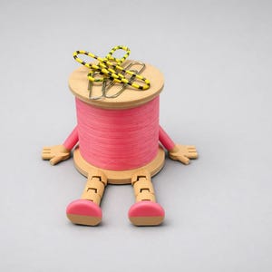 May include: A wooden spool wrapped in bright pink thread, featuring articulated wooden arms and legs with pink feet. The spool has a flat top surface holding a silver metal paperclip and a looped piece of yellow and black patterned cord.
