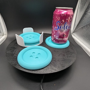 May include: A set of turquoise button-shaped coasters, with one coaster held in a white plastic holder and another coaster supporting a 12 fluid ounce aluminum can of Soleil Black Cherry sparkling water, all arranged on a black circular base.