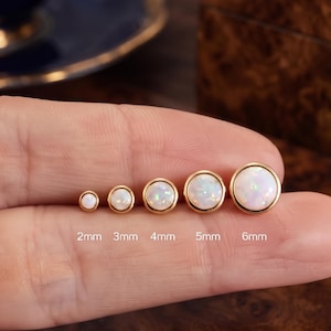 May include: Five round stud earrings with white opals set in gold-toned metal. The earrings are arranged in a row by size, labeled 2mm, 3mm, 4mm, 5mm, and 6mm. Each opal displays iridescent flashes of color within a smooth, polished gold-toned bezel setting.