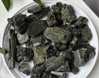 May include: A collection of raw, dark green and black tourmaline crystal shards piled on a round white plate. The stones feature a striated, columnar texture with jagged edges and varying shades of deep olive and charcoal throughout the cluster.
