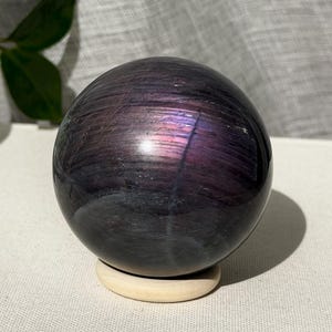 Könnte beinhalten: Ein polierter, kugelförmiger Stein mit einer dunkelvioletten und schwarzen Grundfarbe, der horizontale Bänder mit irisierenden violetten und magentafarbenen Lichtreflexen aufweist und auf einem kleinen, runden Holzständer steht.