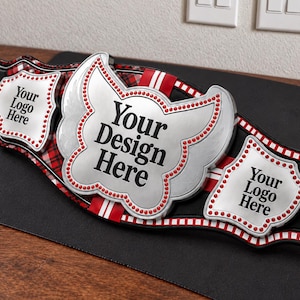 May include: A championship belt featuring a large central silver plate with wings and two smaller side plates. The plates have red beaded borders and the text Your Design Here and Your Logo Here. The strap is black with a red plaid pattern.