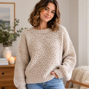 May include: A beige long-sleeve sweater with a textured knit pattern, a round ribbed neckline, and ribbed cuffs. The sweater features a loose fit with dropped shoulders and a thick, chunky knit construction that provides a consistent, uniform appearance.