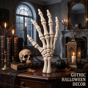 May include: A cream-colored crocheted skeleton hand sculpture standing upright. The hand features detailed finger joints and a wrist base. The text GOTHIC HALLOWEEN DECOR is printed in the bottom right corner of the image.