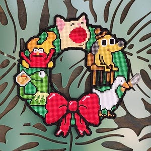 May include: A colorful, handmade wreath featuring cartoon characters, including a pig, dog, frog, and duck. The wreath has a large red bow at the bottom and is set against a green leaf-patterned background.