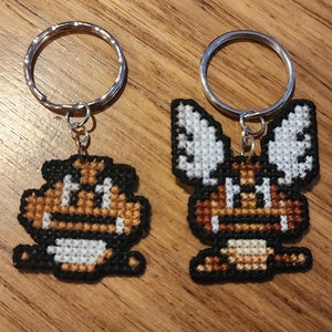 May include: Two pixel art keyrings. One is a brown and black creature with a white belly and mouth. The other is a similar creature with large white and black wings. Both have silver key rings.