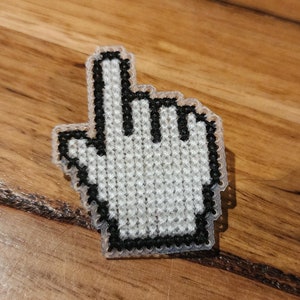 May include: A white pixelated hand cursor with a black outline, pointing upwards. The cursor is made of fabric and is a cross-stitch design.