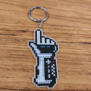 May include: A white and black pixelated keychain with a hand holding a remote control. The hand is pointing up with the index finger extended. The remote control has black buttons and a red button.