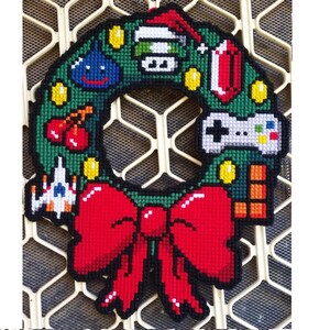 May include: A green and red pixelated Christmas wreath with a red bow, featuring a video game controller, a game cartridge, a Christmas stocking, a cherry, and a pixelated character.