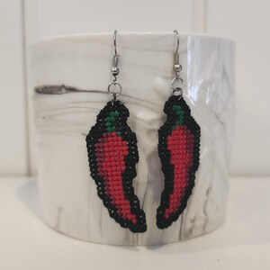Könnte beinhalten: Ein Paar rote Chilischoten-Ohrringe mit schwarzen Umrissen und grünen Spitzen. Die Ohrringe sind aus Stoff und haben silberne Haken.