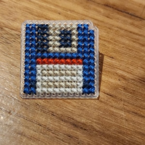 Peut inclure: Une petite broderie au point de croix représentant un disque souple, de couleur bleue, rouge et blanche. La broderie est encadrée dans un cadre en plastique transparent.