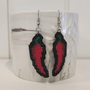 Könnte beinhalten: Ein Paar rote Chilischoten-Ohrringe mit schwarzen Umrissen und grünen Spitzen. Die Ohrringe sind aus Stoff und haben silberne Haken.