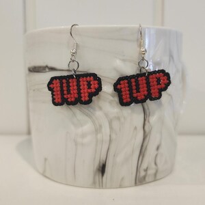 May include: A pair of earrings with red and black pixelated '1UP' charms. The charms are attached to silver hooks.