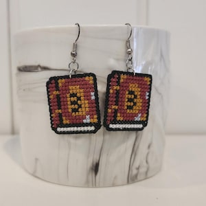 May include: A pair of earrings featuring a cross-stitch design of a closed book with a red cover, yellow pages, and white accents. The earrings are silver and have a hook closure.