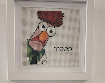 May include: A framed cross-stitch of a red-haired character with large eyes and a green and black shirt. The character is looking to the right and the text "meep" is below the character.