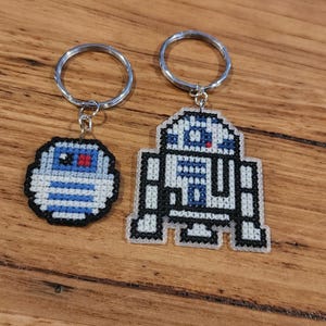 May include: Two keychains featuring pixelated embroidered images of R2-D2 from Star Wars. The larger keychain shows R2-D2 standing, while the smaller keychain shows R2-D2 in a round shape.