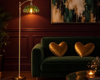 May include: A floor lamp with a slender brass-toned metal stand and a curved arm holding a green, petal-shaped glass shade. The lamp emits a warm, glowing light from an exposed bulb, illuminating the surrounding dark room.