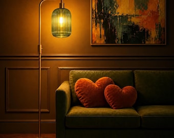May include: A floor lamp with a tall, thin metallic stand and a ribbed, cylindrical green glass shade emitting a warm yellow light. Two plush, heart-shaped orange pillows rest on a dark green sofa positioned next to the lamp.