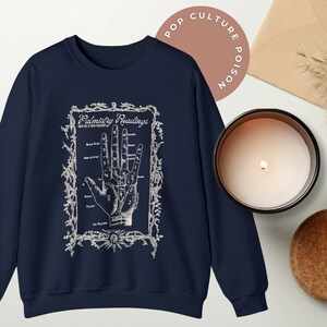 Puede incluir: Sudadera azul marino con un estampado blanco de una mano con l&iacute;neas y marcas etiquetadas como "Palmistry Readings". La impresi&oacute;n est&aacute; rodeada de un marco decorativo.