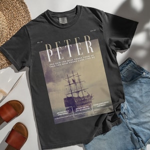 May include: A black t-shirt with a vintage-style graphic featuring a ship sailing on the ocean. The graphic includes the text "PETER", "YOU SAID YOU WERE GONNA GROW UP", "THEN YOU WERE GONNA COME FIND ME", "ARE YOU STILL A MIND READER?", "WE BOTH DID THE BEST WE COULD DO", and "CAUSE LOVE'S NEVER LOST WHEN".