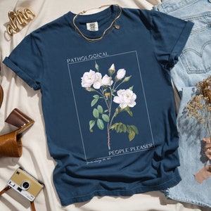 May include: A navy blue t-shirt with a white floral graphic and the text "Pathological People Pleaser".