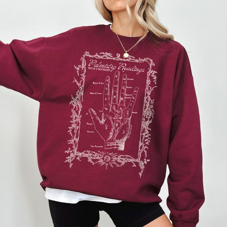 Puede incluir: Sudadera color burdeos con un estampado gr&aacute;fico blanco de una mano con l&iacute;neas y s&iacute;mbolos etiquetados como "Palmistry Readings".