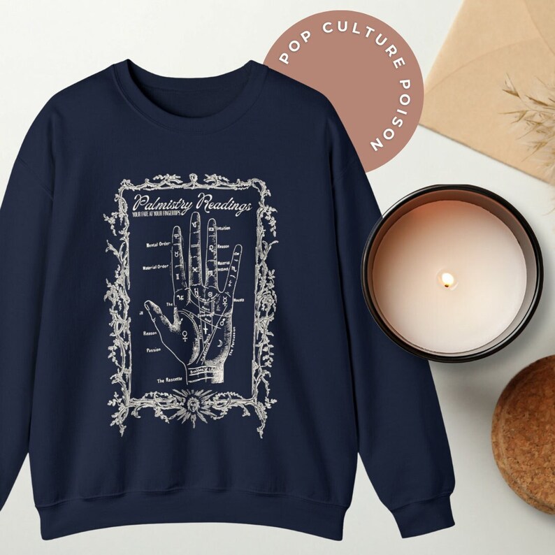 Puede incluir: Sudadera azul marino con un estampado blanco de una mano con l&iacute;neas y marcas etiquetadas como "Palmistry Readings". La impresi&oacute;n est&aacute; rodeada de un marco decorativo.