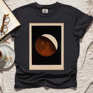 May include: Black t-shirt featuring a vintage-style print of a celestial body, possibly a planet or moon, with a dark background and a light-colored crescent. The print is framed with a beige border. The shirt has a crew neck.