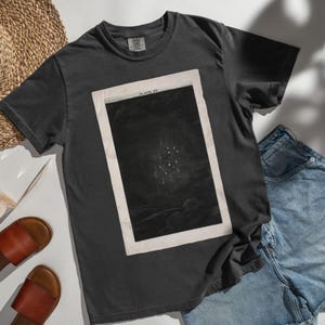 Puede incluir: Una camiseta gris oscuro con una ilustración celestial enmarcada. La obra de arte, etiquetada como "PLATE XV", representa un cúmulo de estrellas sobre un fondo oscuro. La camiseta se combina con pantalones cortos de mezclilla y sandalias marrones, lo que sugiere ropa casual.