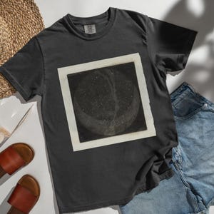 Puede incluir: Camiseta gris oscuro con un diseño abstracto en blanco y negro enmarcado. La obra de arte representa un cuerpo celeste con forma de media luna y detalles moteados. La camiseta se combina con pantalones cortos vaqueros y sandalias de cuero marrón.
