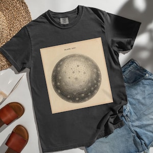 Puede incluir: Camiseta gris oscuro con una ilustración vintage de un cuerpo celeste, etiquetada como "PLATE XXIV". El gráfico es un dibujo circular detallado con numerosos puntos pequeños. La camiseta se combina con pantalones cortos de mezclilla y sandalias de cuero marrón.