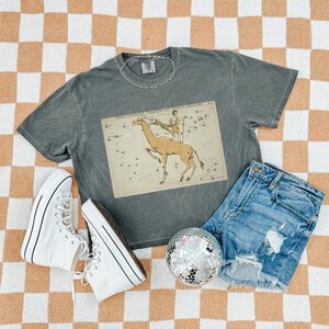 May include: A gray t-shirt with a vintage giraffe constellation graphic, paired with denim shorts, white high-top sneakers, a silver chain necklace, and a disco ball. The outfit is arranged on a checkered orange and white surface.