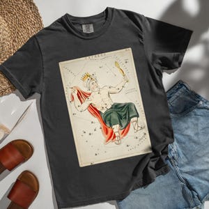 Könnte beinhalten: Dunkelgraues T-Shirt mit einer Vintage-Illustration des Sternbilds Cepheus. Das Kunstwerk zeigt eine gekrönte Figur mit einem Zepter, rotem Umhang und grüner Kleidung. Das T-Shirt ist ein bequemer, lässiger Stil.